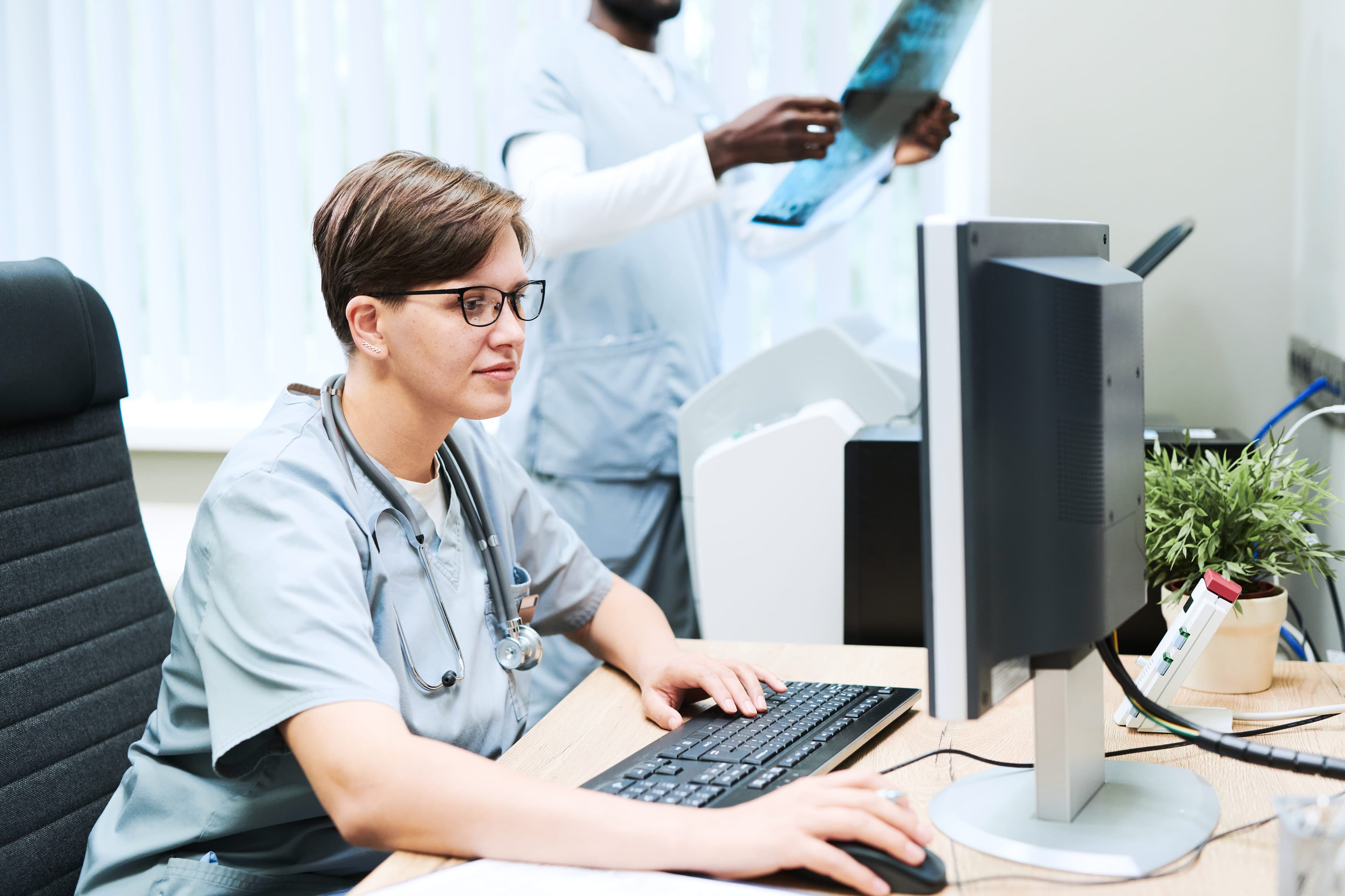 A clinician working at a computer while a colleague reviews an X-ray behind her