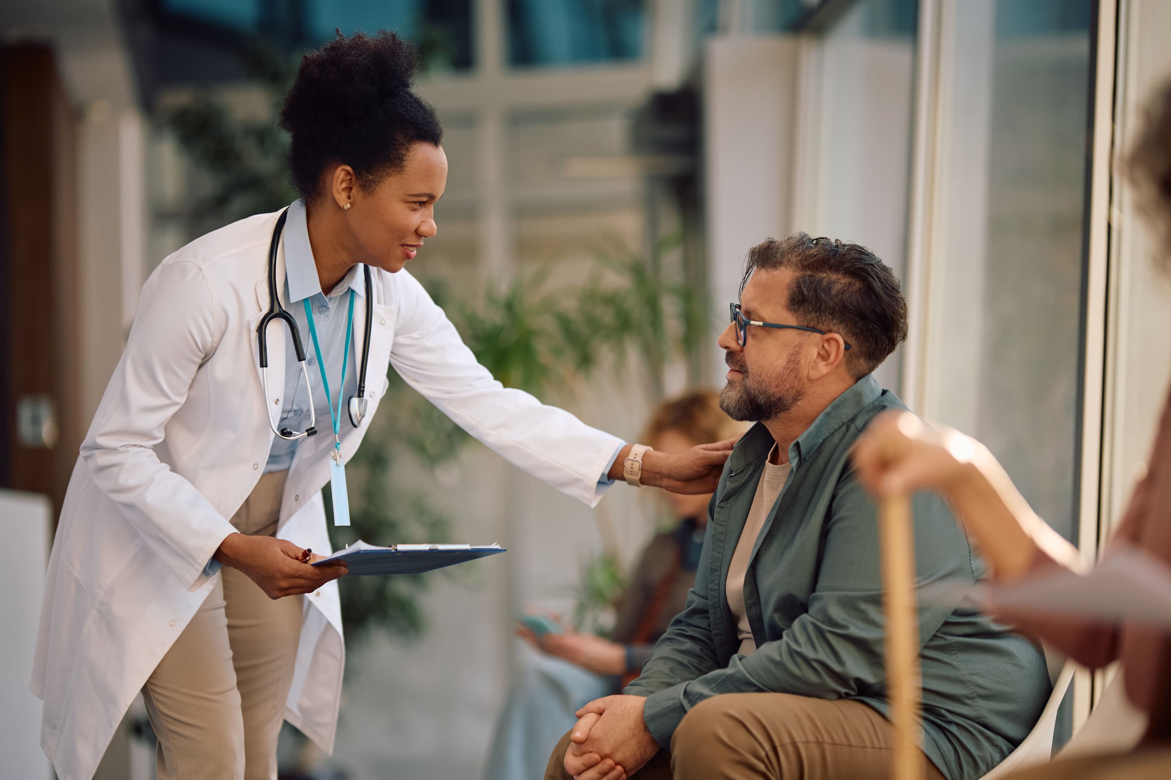 A physician with a clipboard leaning in to talk with a seated patient